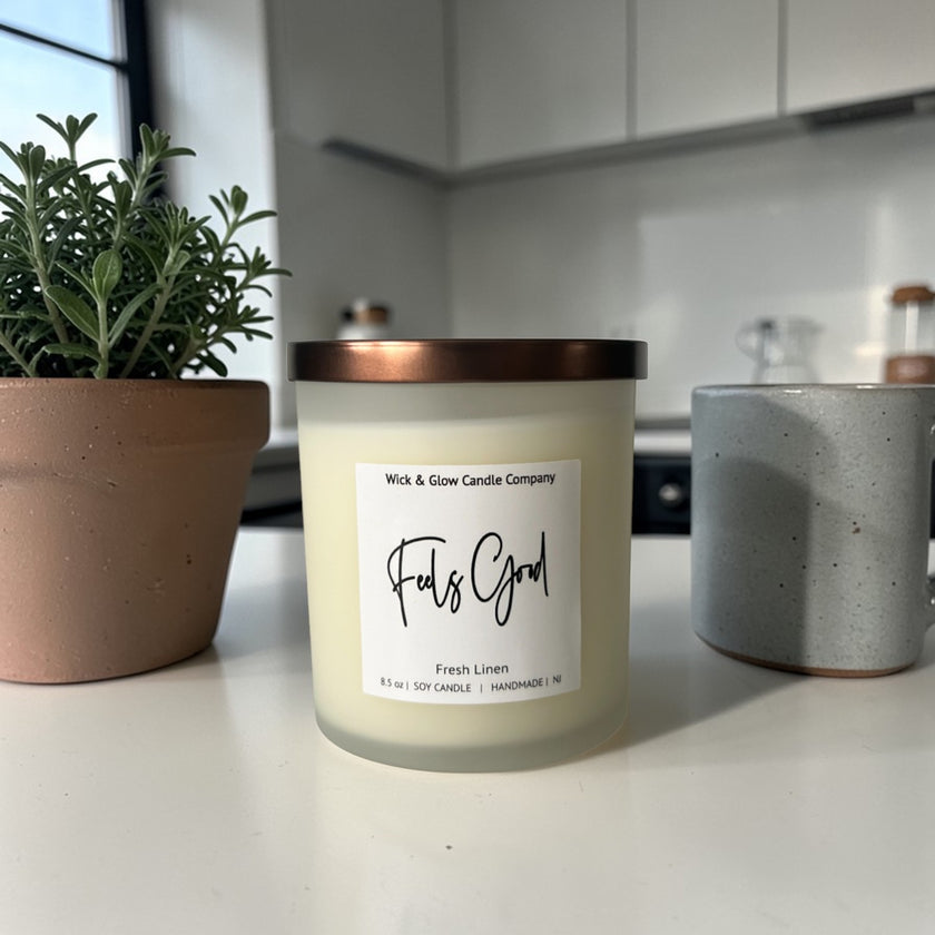 Candle labeled 'Feels Good' on a kitchen counter with a plant and container in the background