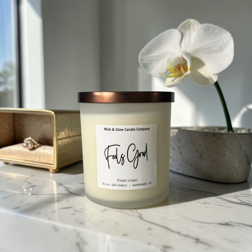 Candle labeled 'Feels Good' by Wick & Glow Candle Company on a marble surface with a white orchid in the background.