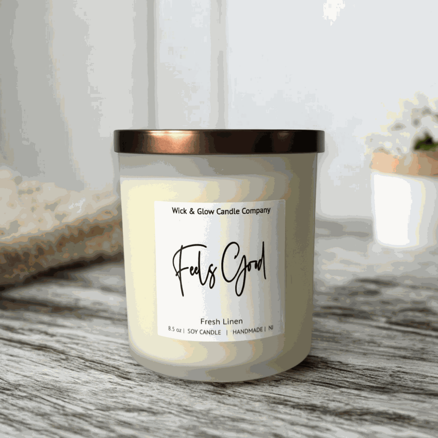 A  fresh linen soy wax candle in a frosted jar with a bronze lid from Wick and Glow Candle Company on agreyable with plants in the background