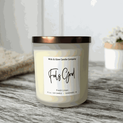 A  fresh linen soy wax candle in a frosted jar with a bronze lid from Wick and Glow Candle Company on agreyable with plants in the background