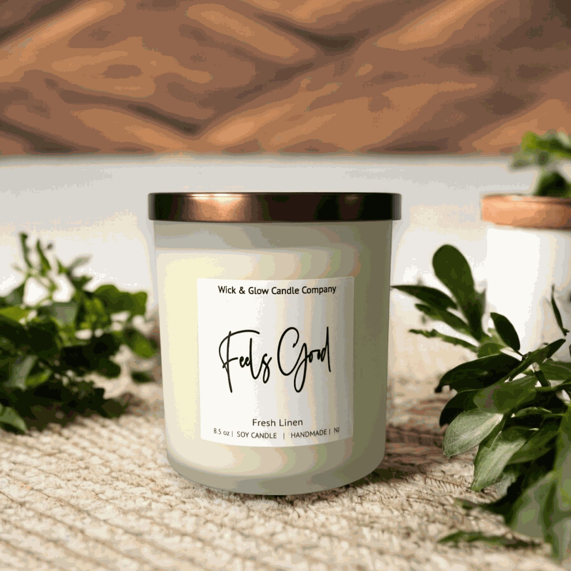 A fresh linen soy wax candle in a frosted jar with a bronze lid from Wick and Glow Candle Company on a brown table with plants in the background.