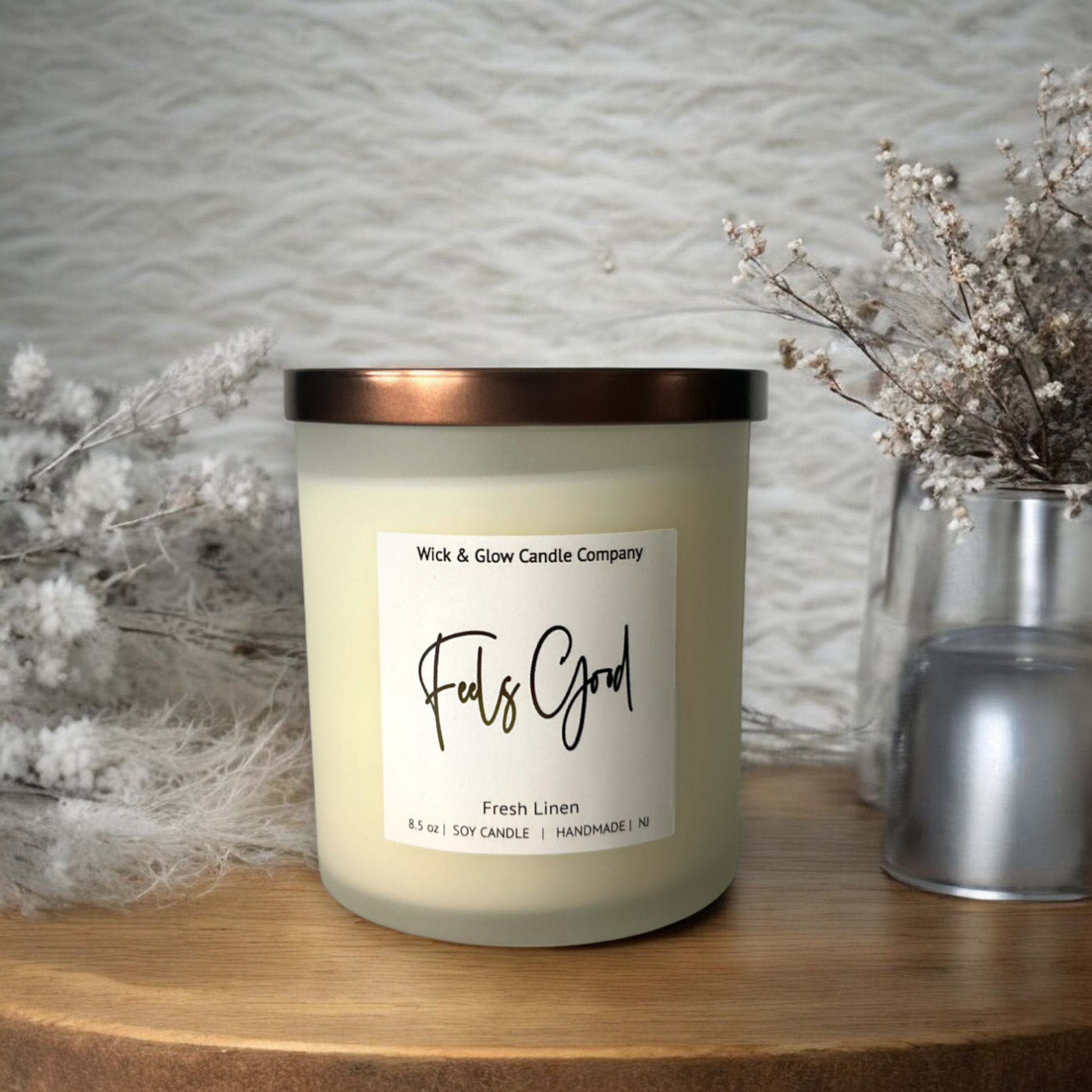 A soy wax candle in a frosted white jar with bronze lid displayed on a rustic wooden table, with white flowers.