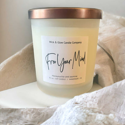 A white candle in a frosted glass jar with a bronze lid with a white label on a wooden table with a white background from Wick and Glow Candle Company.