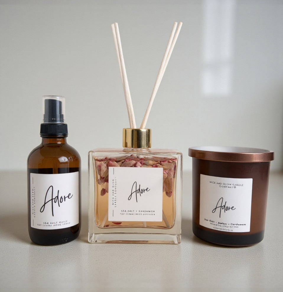 Three Adore products including a bottle, diffuser, and candle on a light surface.