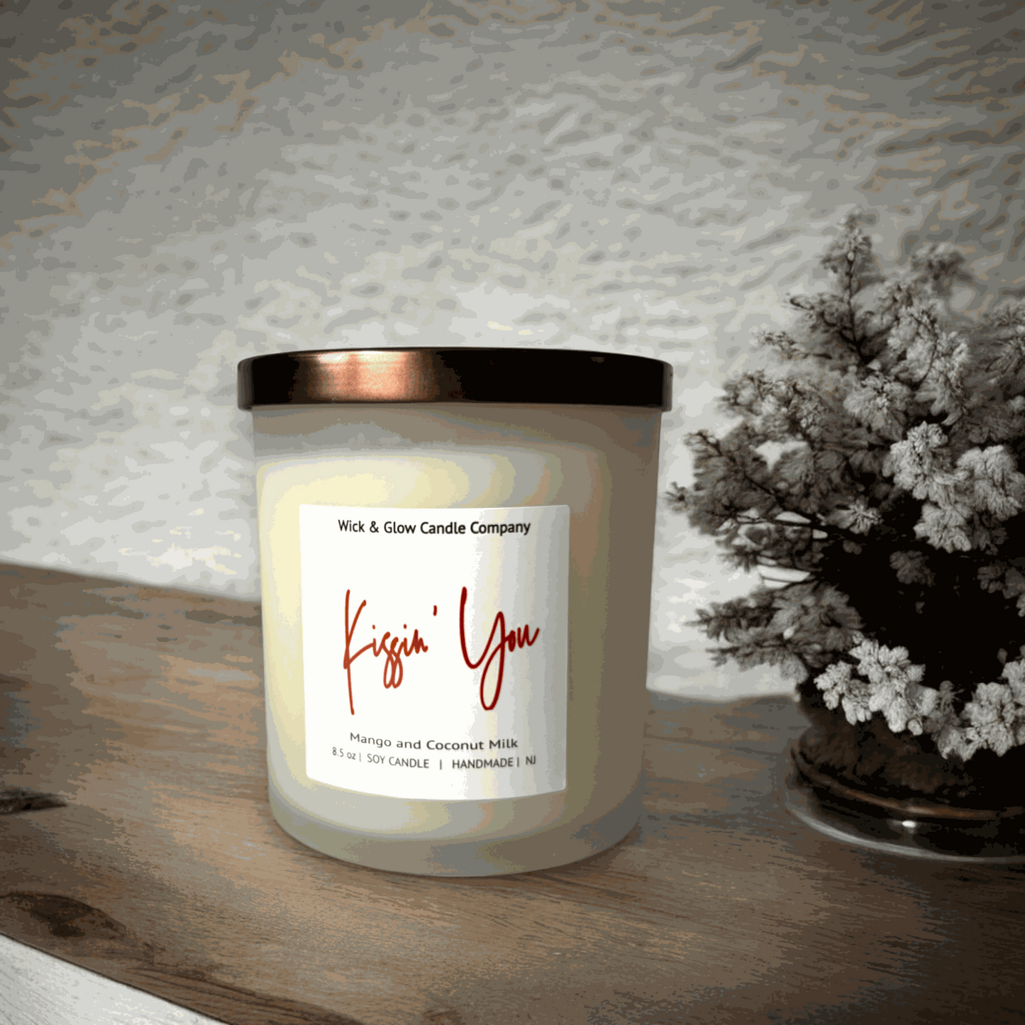 A white candle in a frosted glass jar with a bronze lid with a white label  from Wick and Glow Candle Company on a wooden table with a plant in the background.