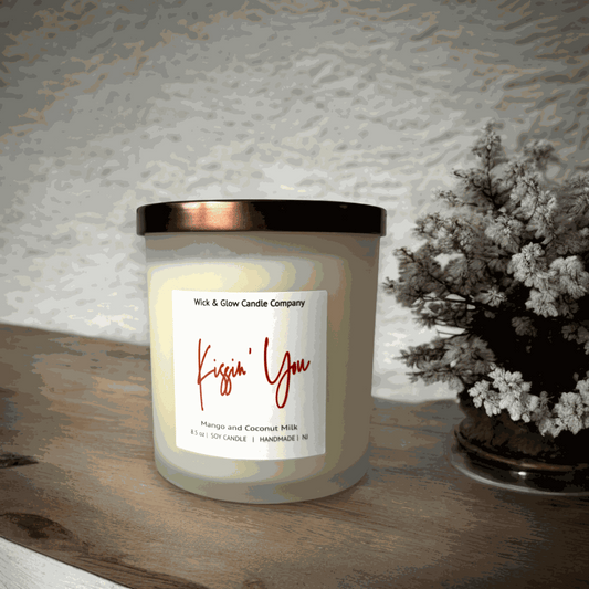 A white candle in a frosted glass jar with a bronze lid with a white label  from Wick and Glow Candle Company on a wooden table with a plant in the background.