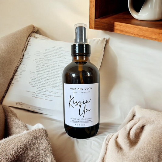 Amber spray bottle  with a white label from the Wick and Glow Candle Company on a white table with white and tan blankets in the background ; the scent is mango and coconutmilk