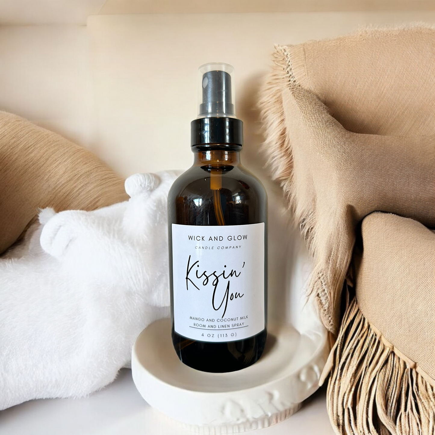 Amber spray bottle  with a white label from the Wick and Glow Candle Company on a white table with white and tan blankets in the background ; the scent is mango and coconutmilk