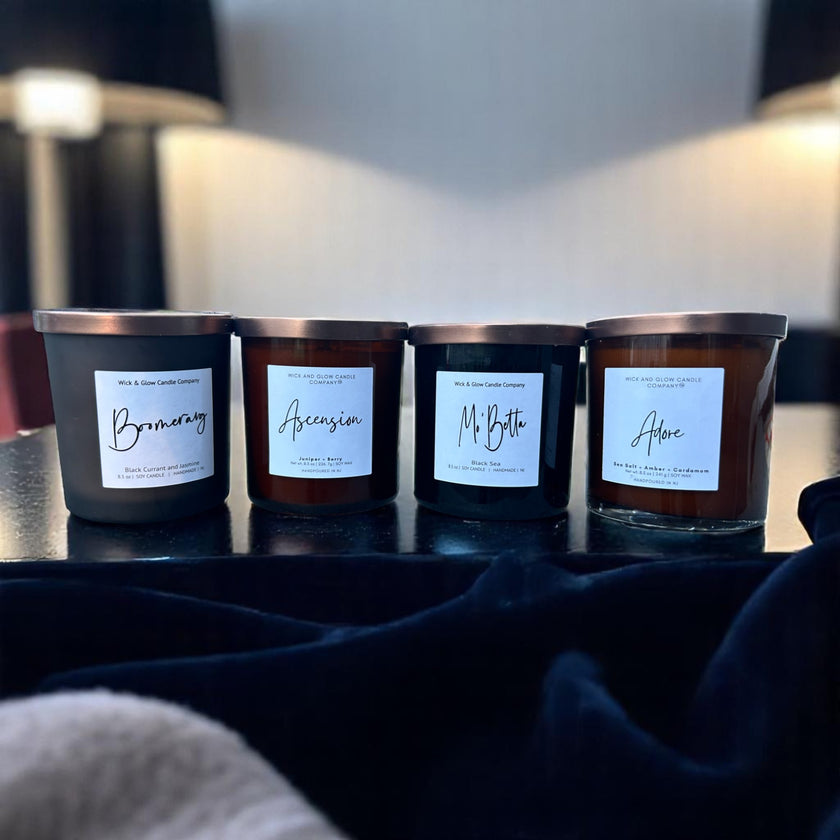 A set of four soy wax candles with labels in a line, each in a black container with different scents indicated by the labels Boomerang, Adore, Ascension, and Mo Betta.