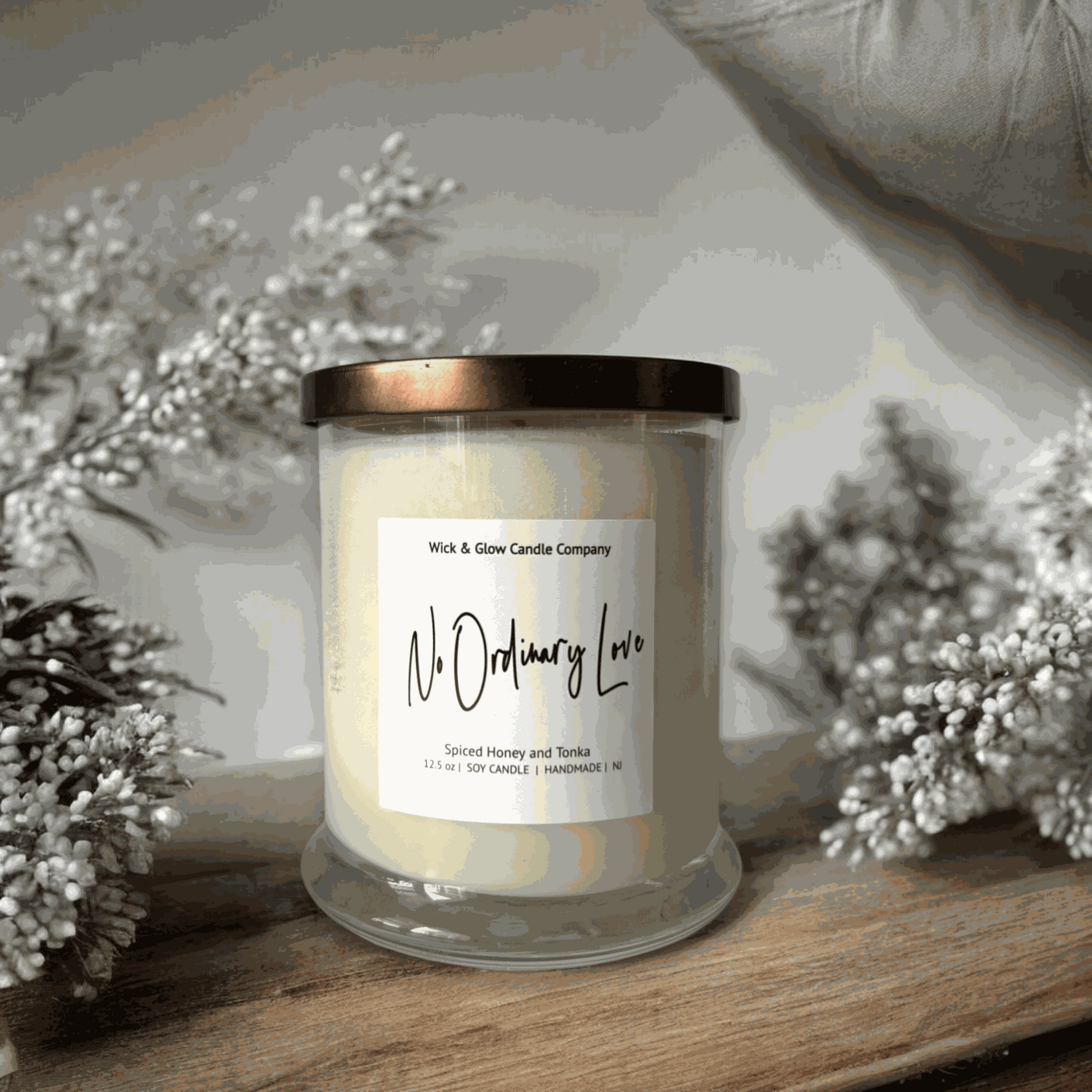 A candle in a clear jar with bronze lid and white label from Wick and Glow Candle Company on a wooden table with a white background and white branches. Scents is Spiced Honey and Tonka