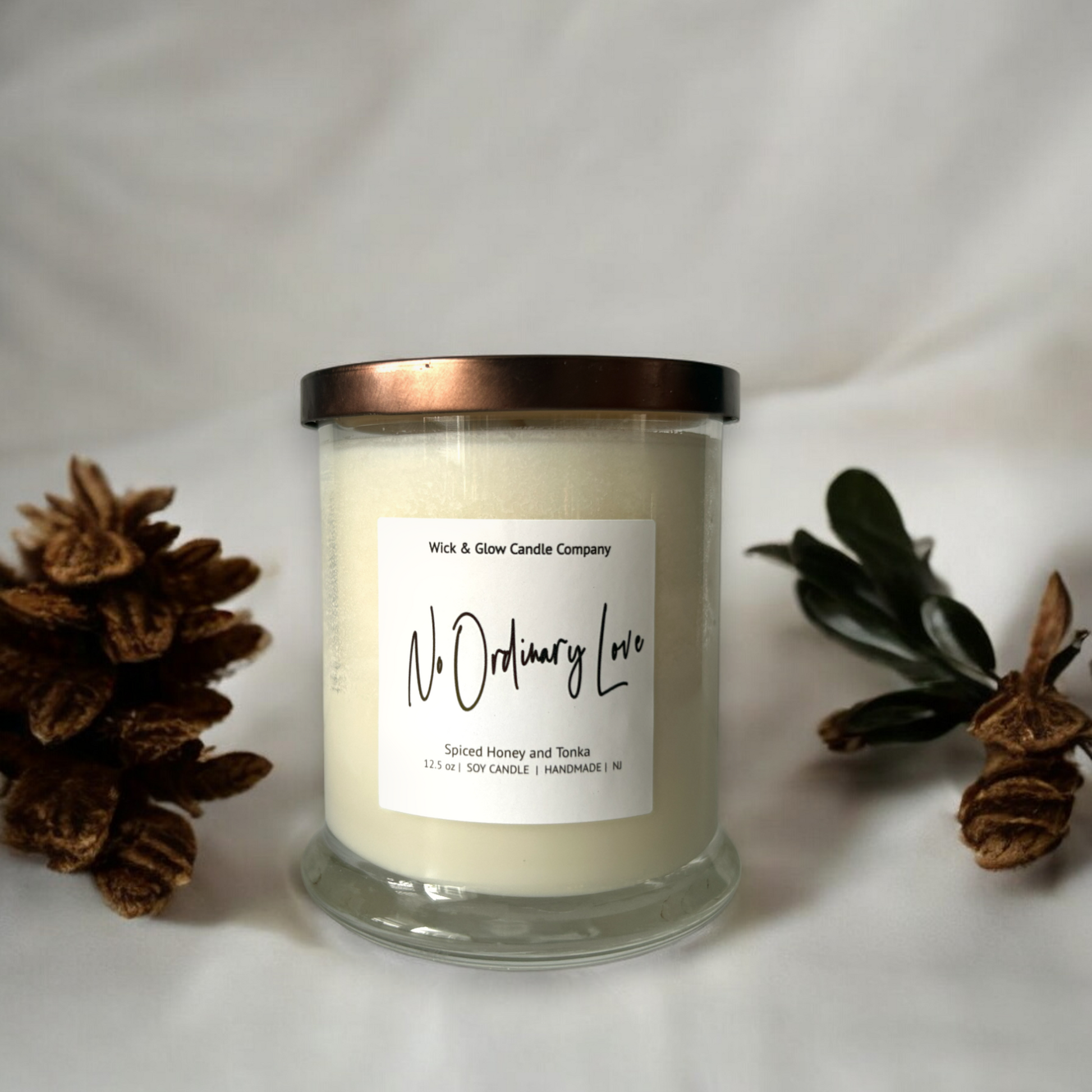 White candle in a clear jar with a bronze lid on a white background with pine cones. The scent is spiced honey and tonka.