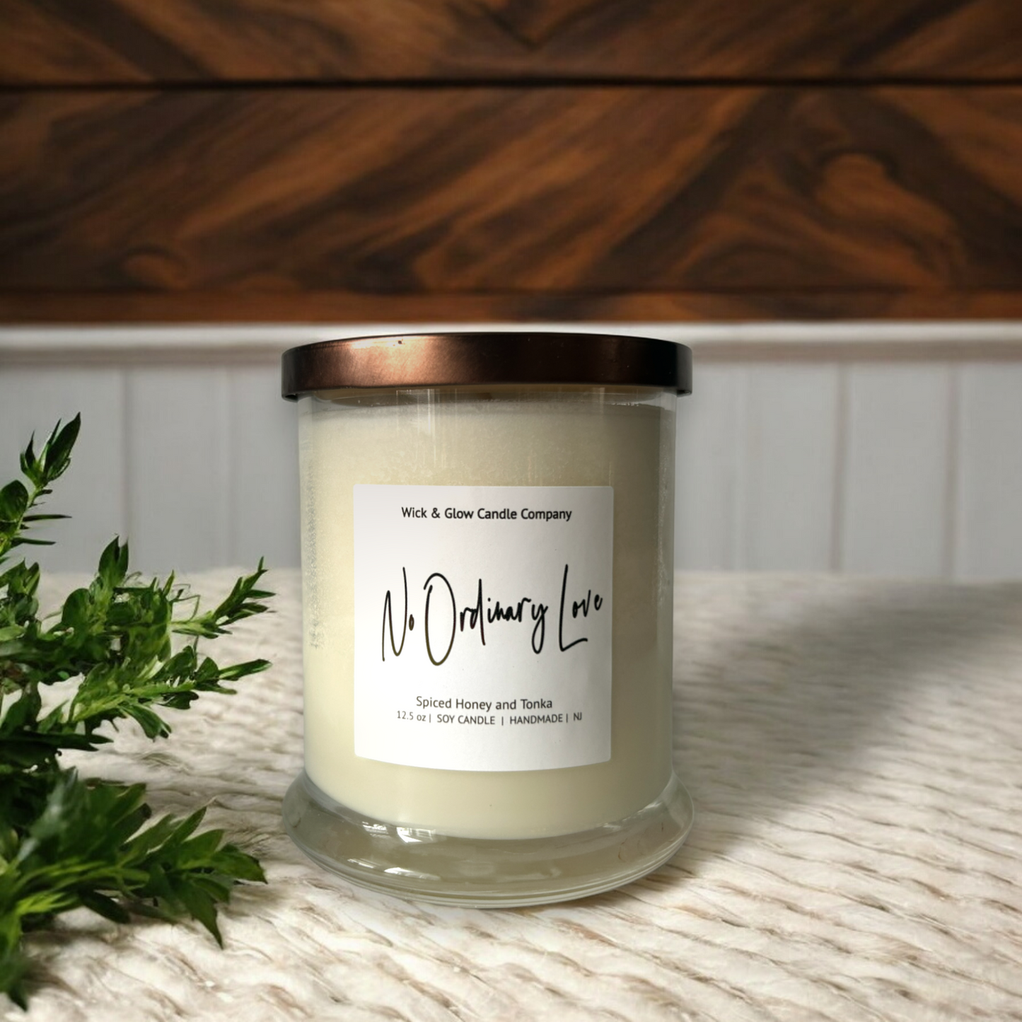 White candle in a clear jar with a bronze lid on a tan table cloth with a wooden and white background. The scent is spiced honey and tonka