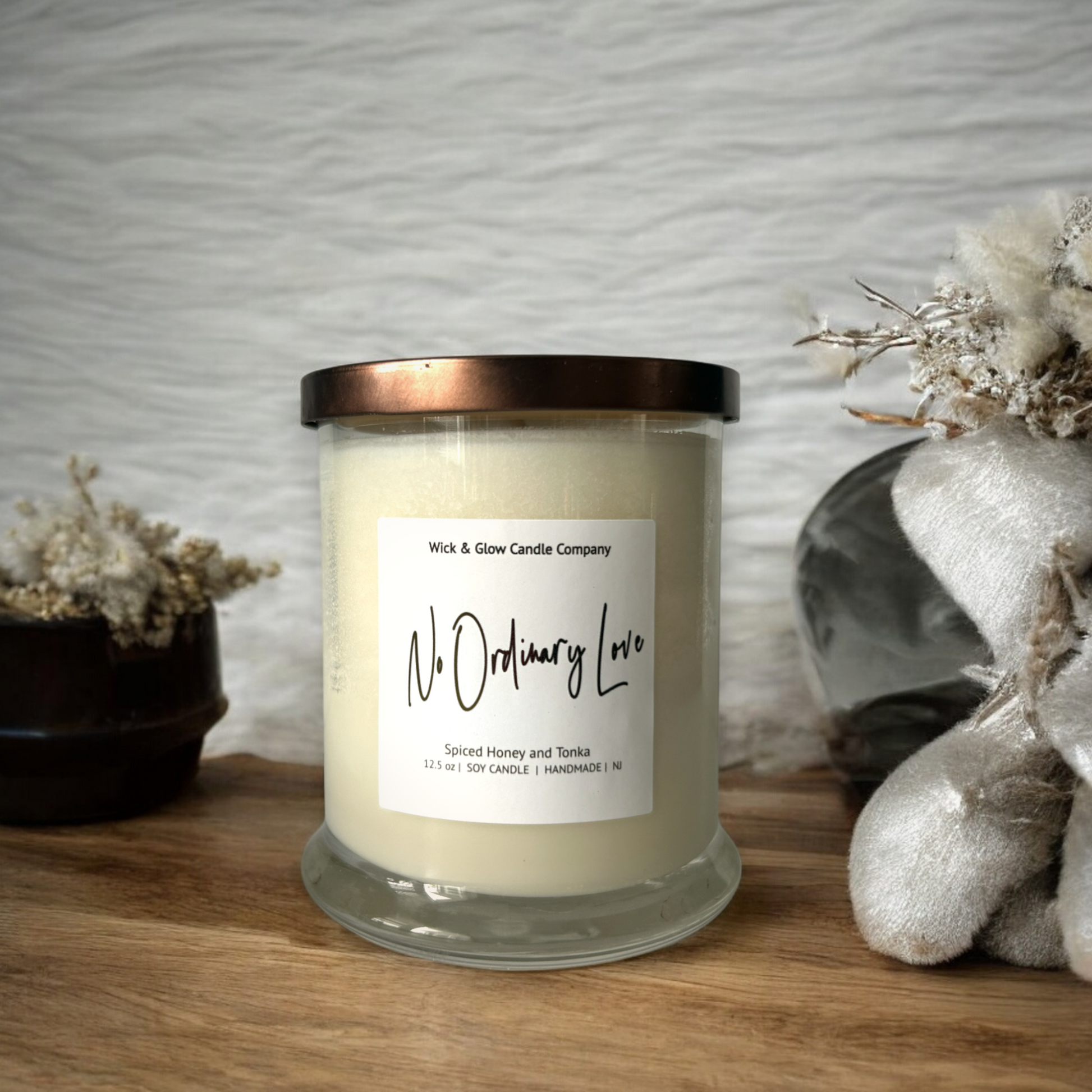 White candle in a clear jar with a bronze lid on a wooden table surrounded by white plants. The scent is spiced honey and tonka.