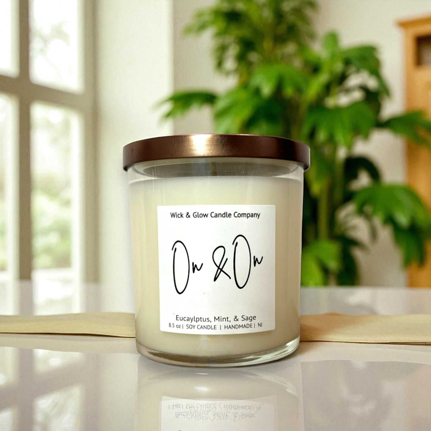 Candle with a label on a reflective surface, blurred plant in the background with a Wick and Glow label