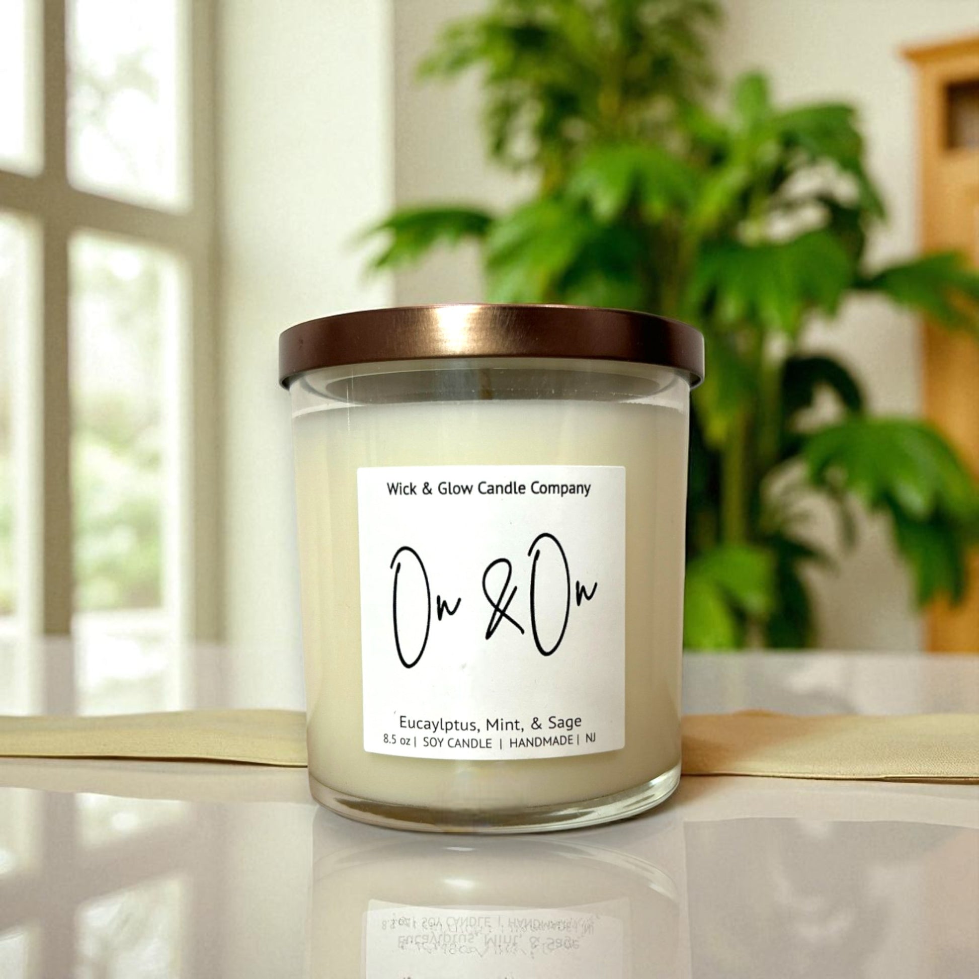 Candle with a label on a reflective surface, blurred plant in the background with a Wick and Glow label