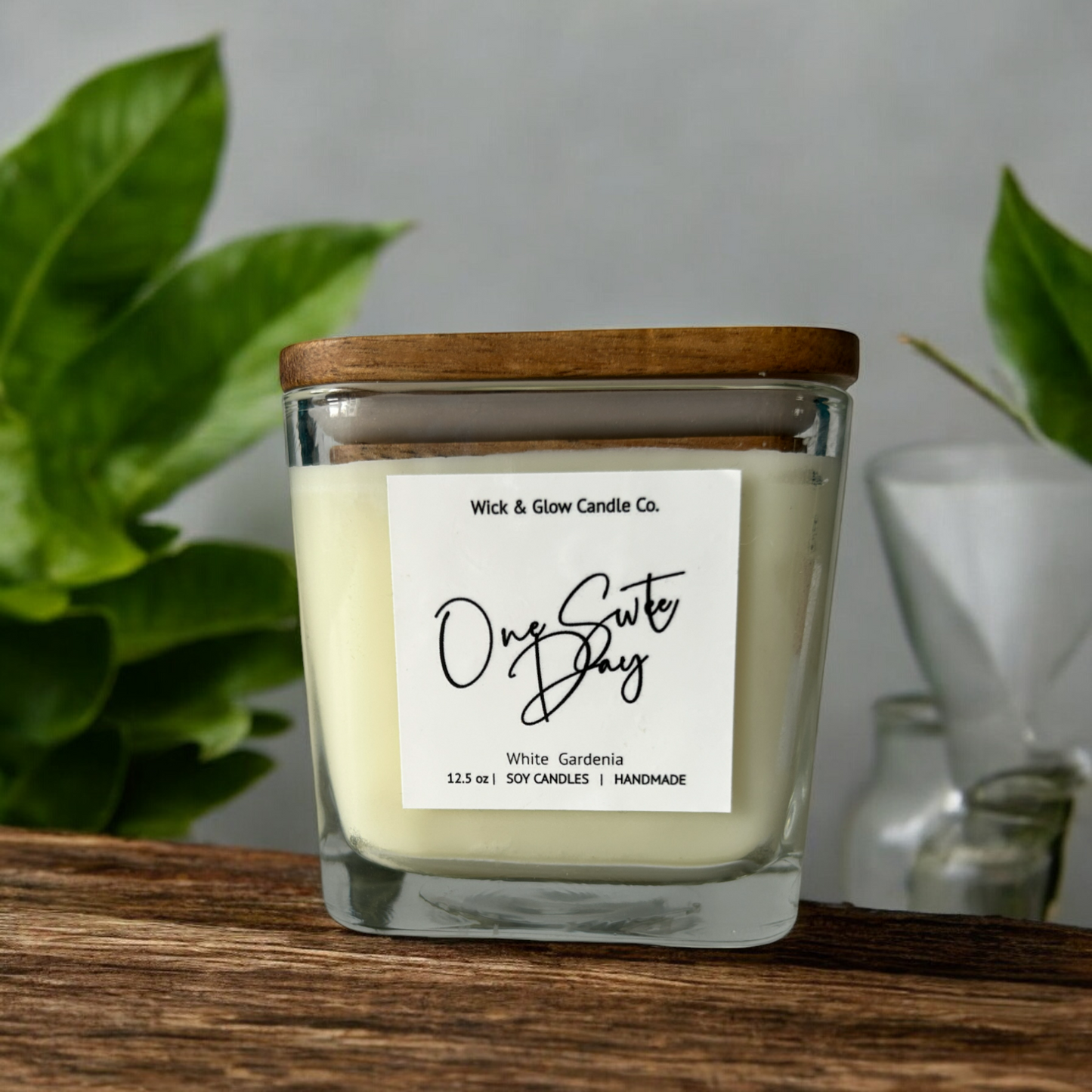 White candle in a clear square jar with a wooden lid on a wooden table  with white background surrounded by plants.