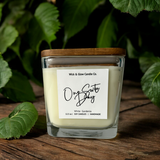 White candle in a clear square jar with a wooden lid on a wooden table surrounded by plants.