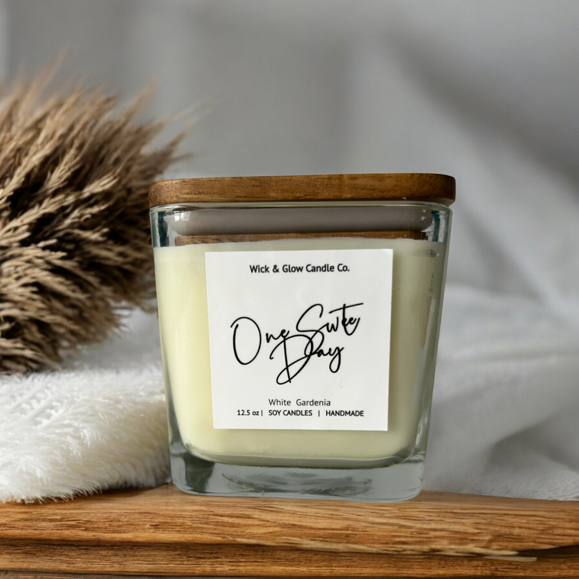 White candle in a clear square jar with a wooden lid on a wooden table  with a white background  and a brown plant.