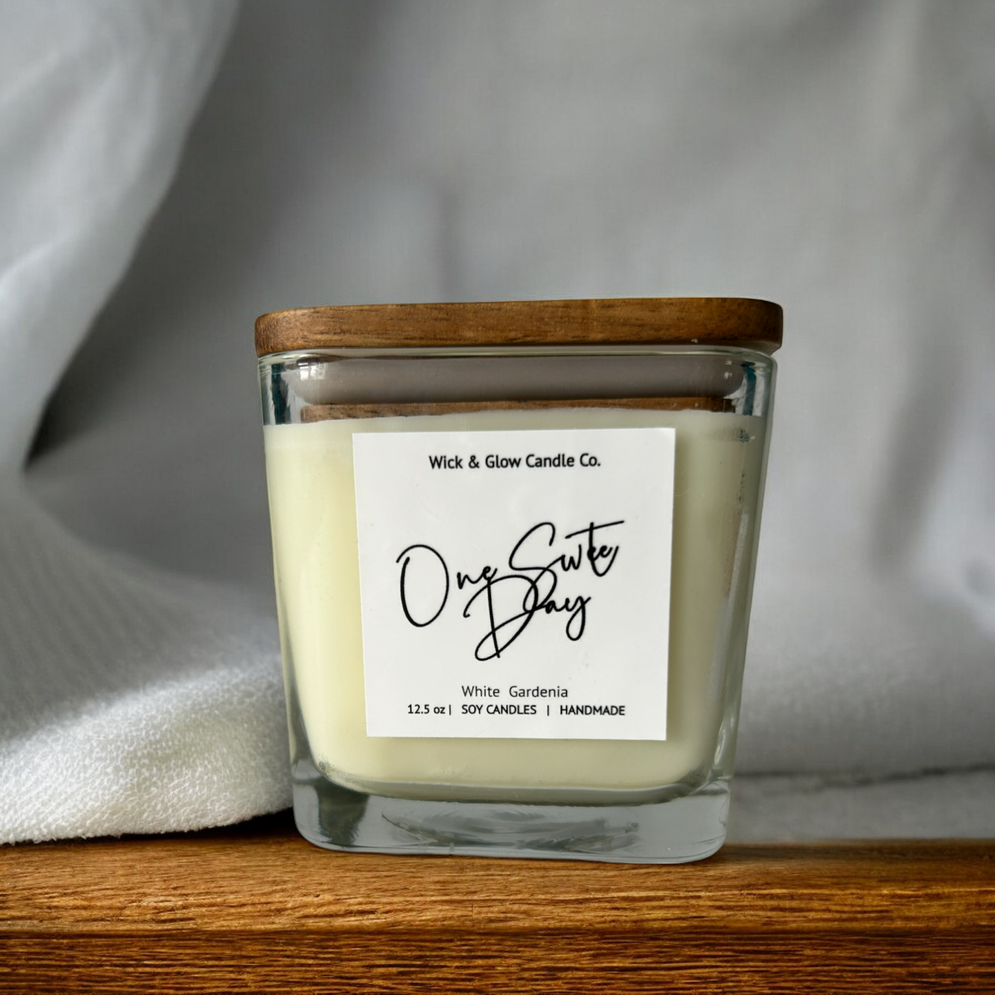 White candle in a clear square jar with a wooden lid on a wooden table with a white background.