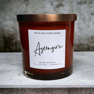 Candle in a brown jar with a bronze lif on a concrete table with a white label that reads Ascension