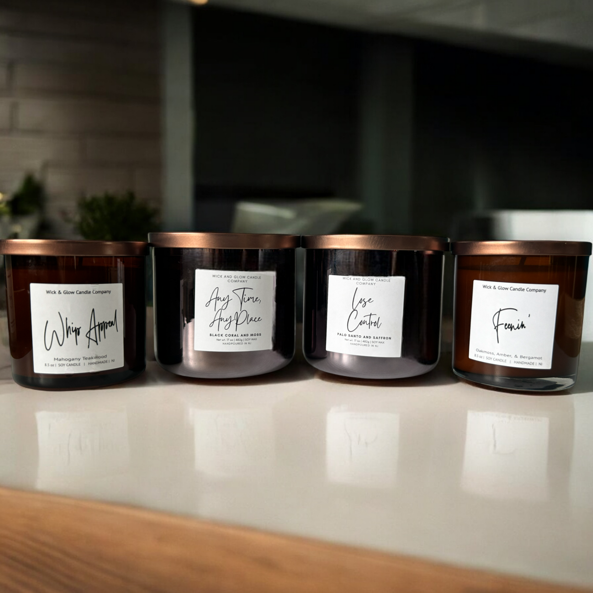 4 candles in black and amber jars with bronze lids sitting on a white table with a living room in the background. The scents are Mahogany and Teakwood, Black Coral and Moss, Palo Santo and Saffron and Oakmoss Amber and Bergamot.