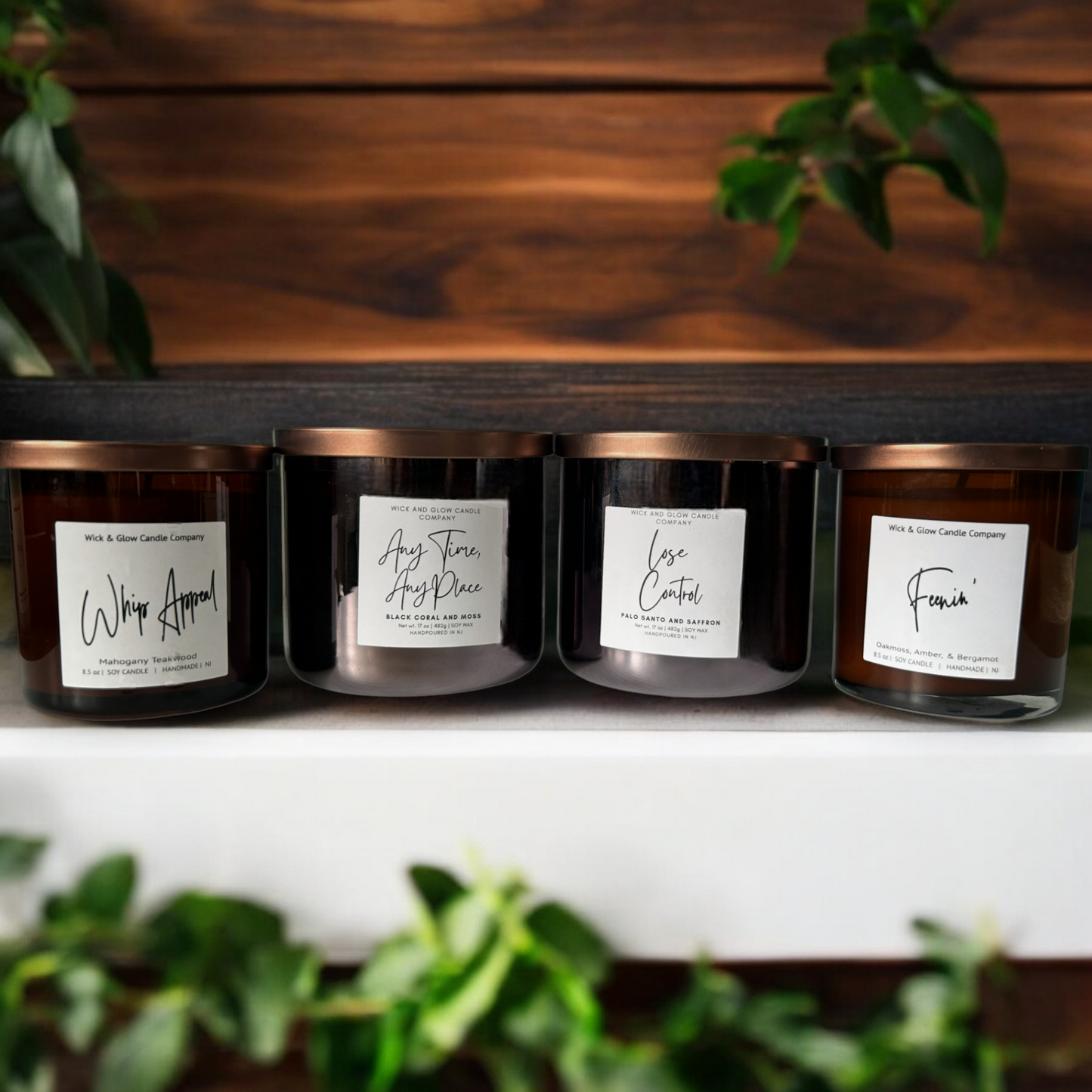 4 candles in black and amber jars with bronze lids sitting on a white table with a living room in the background. The scents are Mahogany and Teakwood, Black Coral and Moss, Palo Santo and Saffron and Oakmoss Amber and Bergamot.