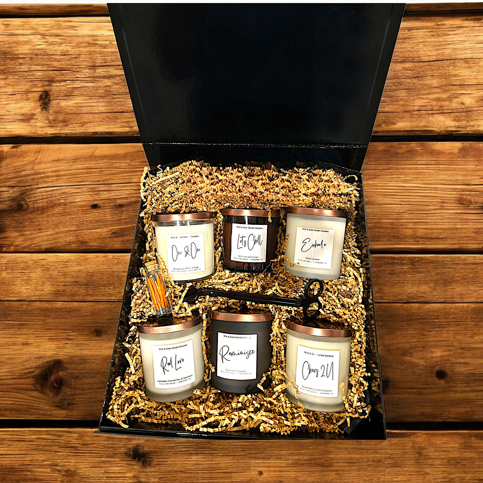 Welcome Home Box black gift box with six candles in black and white glass jars and a wick cutter and matches in brown craft paper on a wooden table.