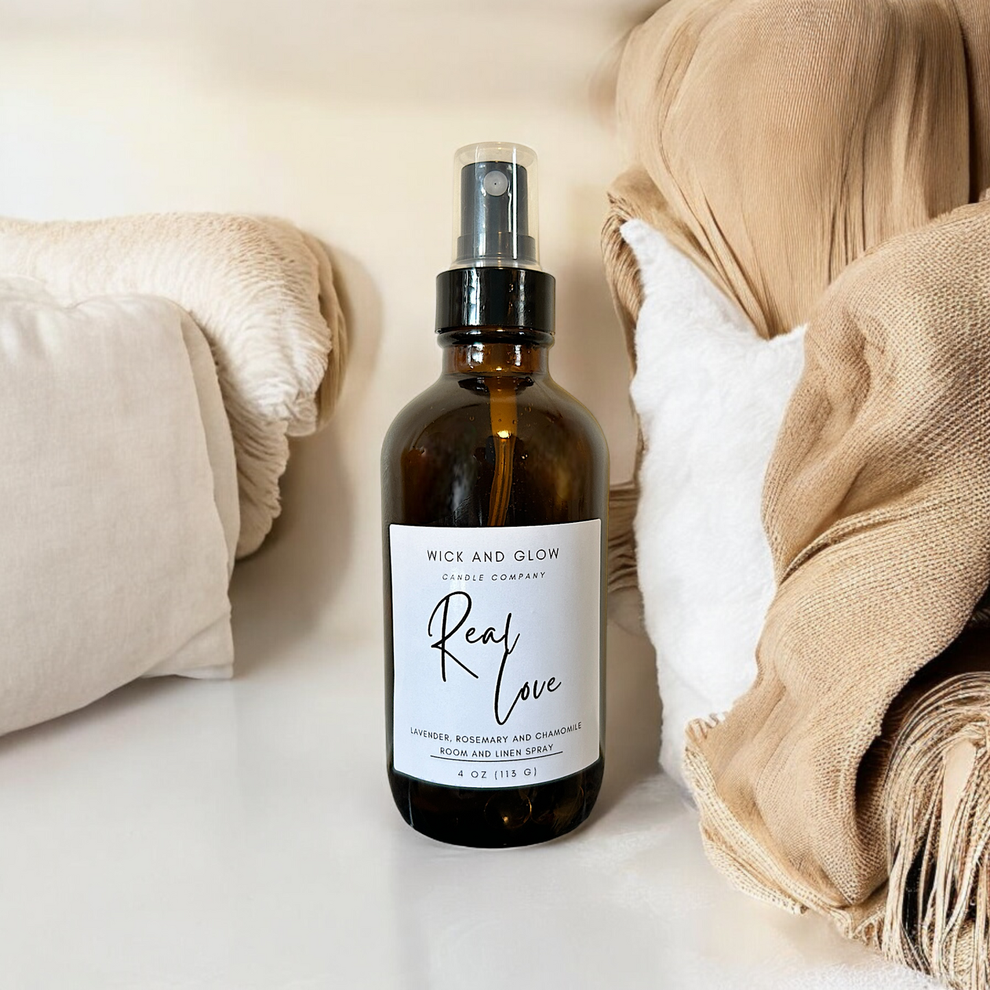 Amber spray bottle  with a white label from the Wick and Glow Candle Company on a white table with tan blankets and white pillows in the background ; the scent is Lavender, Rosemary and Chamomile