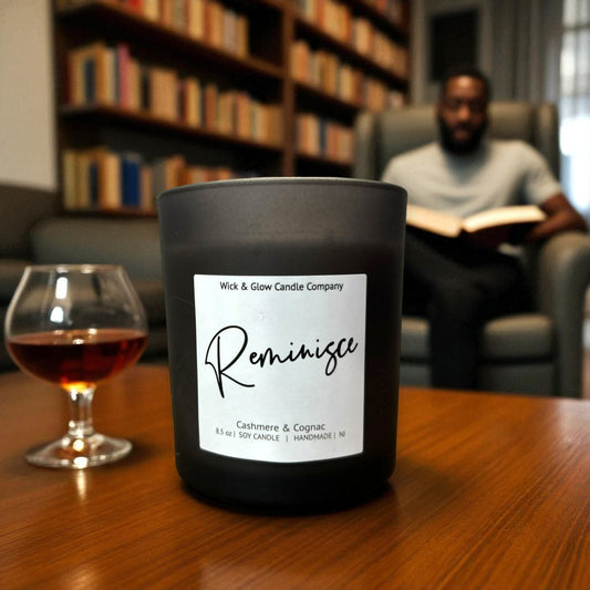 Black candle labeled 'Reminisce' on a table with a glass of whiskey and a person reading in the background.