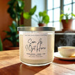 Candle labeled 'Soon As I Get Home' by Wick and Glow Candle Company on a wooden table with plants in the background.