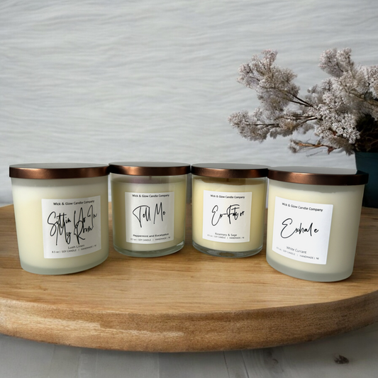 4 candles in clear jars with bronze lids on a wooden table surrounded by white plants on a white background.