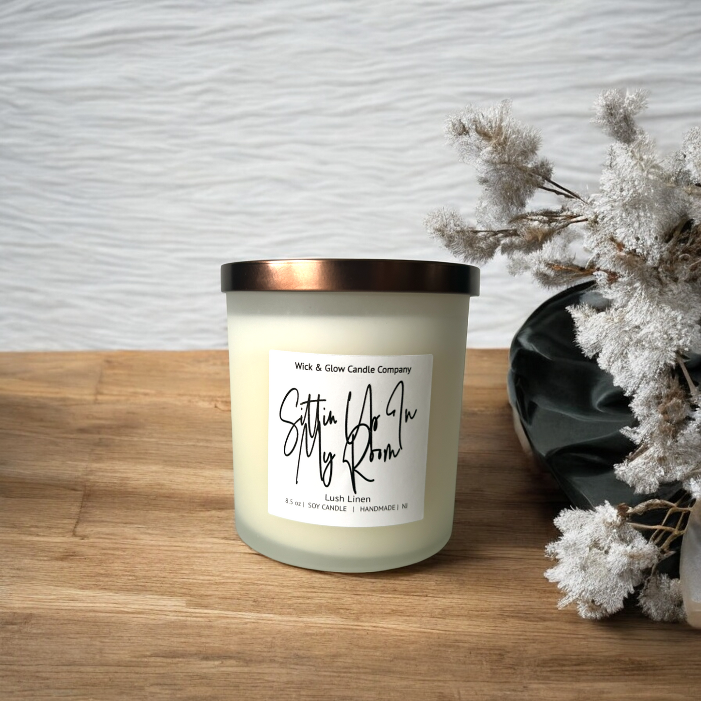 White candle in a clear jar with a bronze lid on a wooden table surrounded by white plants. The scent is lush linen