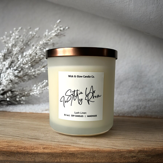 A luxury scented candle in a clear jar with a bronze lid sitting on a wooden table with white flowers in the background.