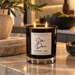 A lit double-wick candle in a clear glass jar sits on a marble countertop in a warm, modern kitchen setting. The candle features a white label with black text that reads 'WICK AND GLOW CANDLE COMPANY' and 'Slow Down CASHMERE + MUSK'.