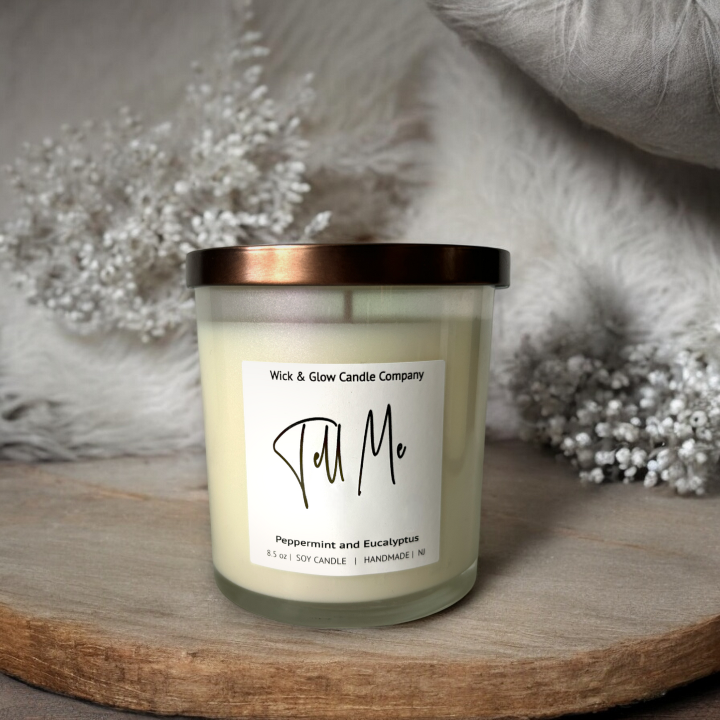 White candle in a clear jar with a bronze lid on a wooden table surrounded by white plants. The scent is peppermint and eucalyptus.