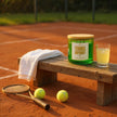 Candle labeled 'Tennis Anyone?' on a bench with tennis balls and a racket on a clay court.