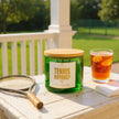 Candle labeled 'Tennis Anyone?' on a table with a tennis racket and iced tea.