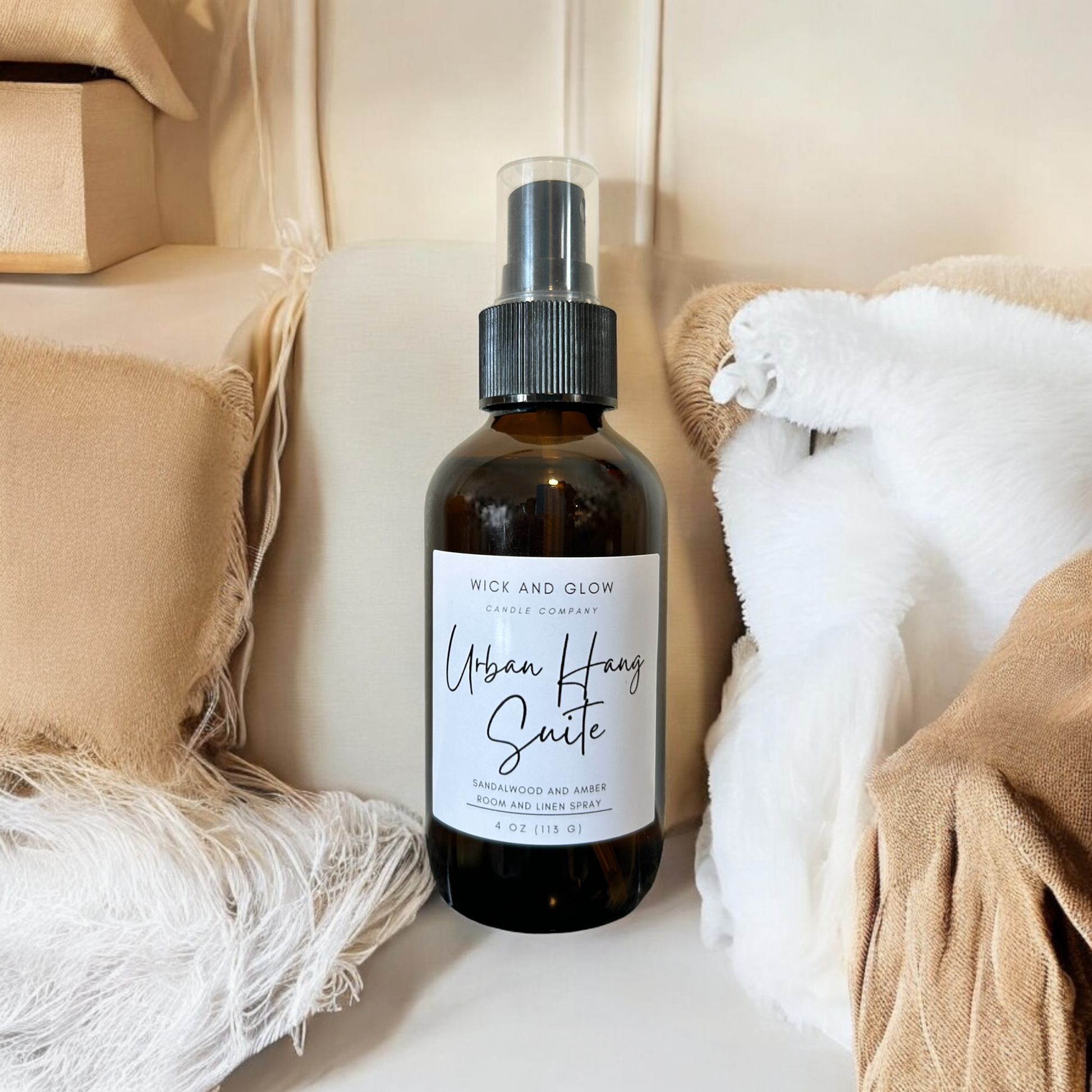 Amber spray bottle with a white label from the Wick and Glow Candle Company on a white table with a white in the background with tan and white pillows ; the scent is Sandalwood and Amber