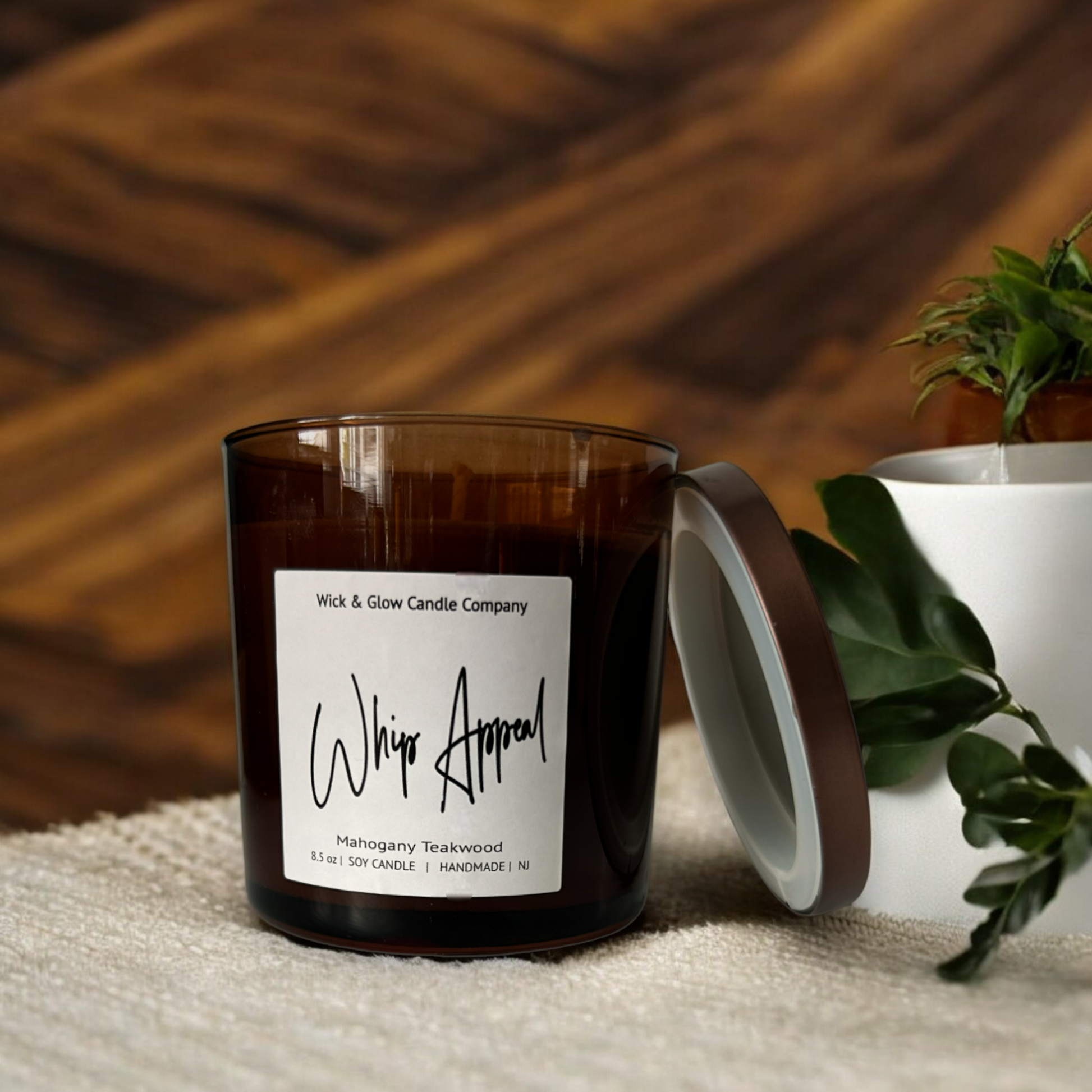 Candle in an amber jar on a tan tablecloth on a wooden background.  The scent is Mahogany Teakwood.