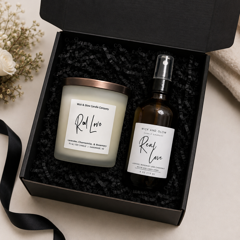 A black gift box with shredded black paper insert holding a white wax candle and an amber spray bottle, both labeled Rice and Glow Candle Company Real Love. The box is open on a light beige surface with a black ribbon and small dried flowers nearby.