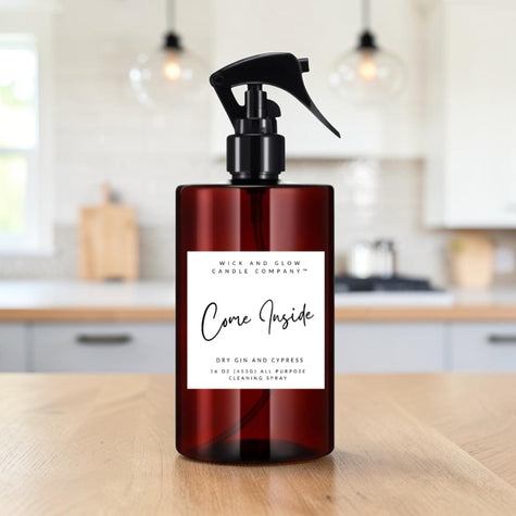 A dark amber spray bottle with a black trigger top and a white label reading 'Come Inside' by Mice and Glow Candle Company, placed on a wooden table with a softly blurred kitchen background.
