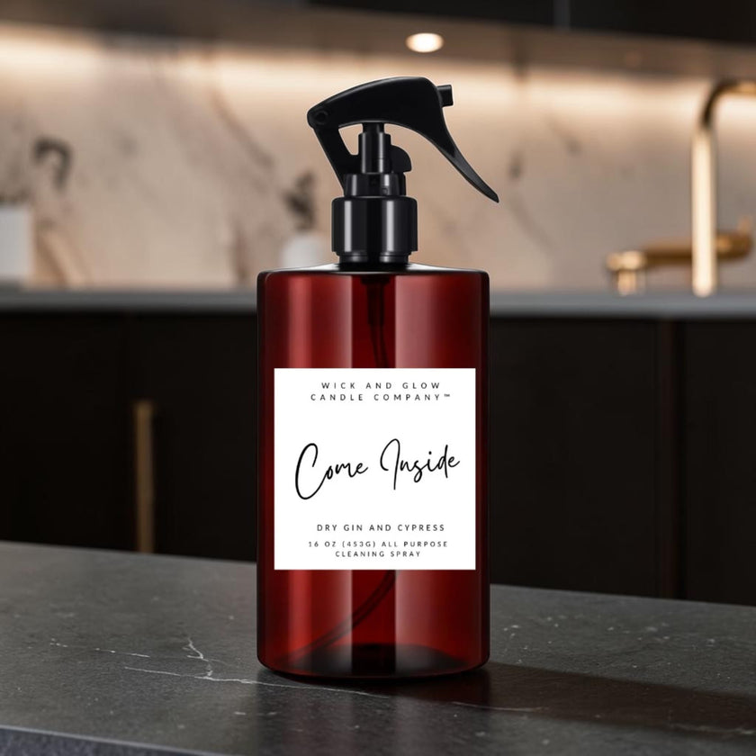 A dark amber glass cleaning spray bottle with a black trigger sprayer and a white label reading 'Come Inside' by Mice and Glow Candle Company, set on a dark countertop with a softly blurred kitchen background.