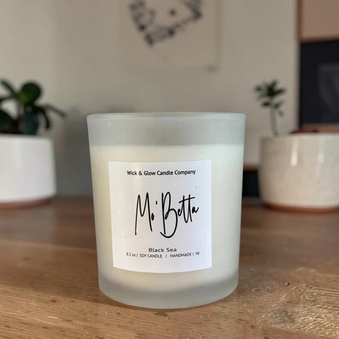 A frosted glass jar candle labeled 'Mr. Botta' by Wick & Glow Candle Company, placed on a wooden surface with potted plants in the blurred background.