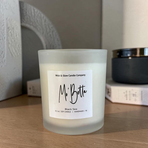 A frosted glass jar candle labeled 'Mr. Botte' by Wick & Glow Candle Company, placed on a wooden surface with books and a dark container blurred in the background.